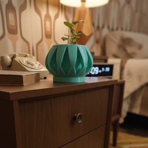 May include: A teal-colored, geometric-patterned planter with a small plant inside. The planter sits on a wooden nightstand with a drawer and a round metal knob. A vintage rotary phone and a book are also on the nightstand.