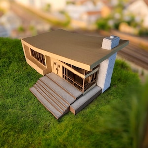 May include: A miniature model of a modern house with a brown roof, white chimney, and a set of stairs leading to the front door. The house is sitting on a bed of green grass.
