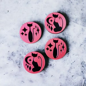 May include: Four pink round coasters with a black silhouette of a cat sitting with a crescent moon and stars behind it.