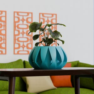May include: A teal-colored, geometric planter with a leafy green plant sits on a dark brown table. The planter has a unique, faceted design. In the background, a green sofa and orange wall decor add a pop of color to the room.