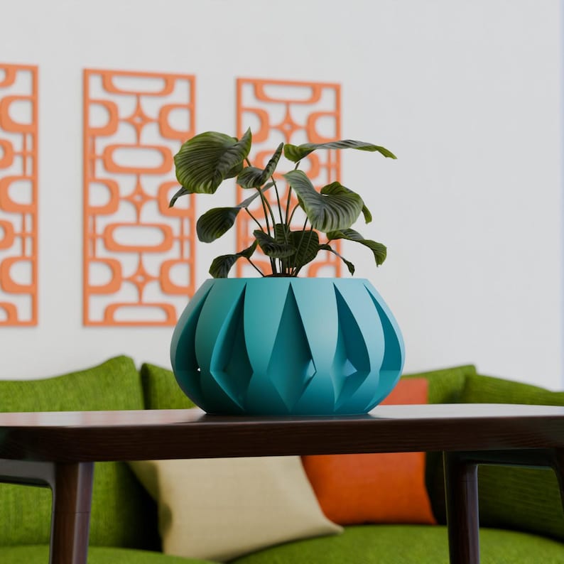 May include: A teal-colored, geometric planter with a leafy green plant sits on a dark brown table. The planter has a unique, faceted design. In the background, a green sofa and orange wall decor add a pop of color to the room.