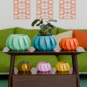 May include: A collection of colorful, geometric planters in shades of green, blue, orange, gold, pink, and yellow. One planter holds a small plant. The planters are arranged on a dark wooden table, with a green sofa and orange pillows in the background.