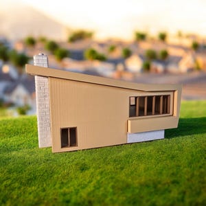 May include: A beige miniature model of a modern house with a flat roof and a chimney. The house has a large window on the side and a smaller window on the front. The model is sitting on a green grassy surface.