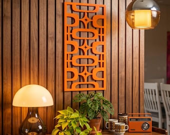 May include: An orange mid-century modern wall panel with a geometric design is mounted on a wood-paneled wall. A table displays a lamp, plants, mugs, and an orange radio. A pendant light hangs from the ceiling.