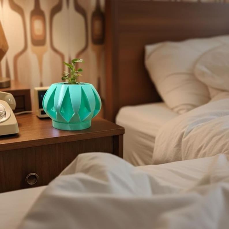 May include: A teal-colored, geometric-patterned planter with a small green plant inside. The planter sits on a dark brown nightstand next to a white telephone and a digital clock. The background includes a bed with white bedding.