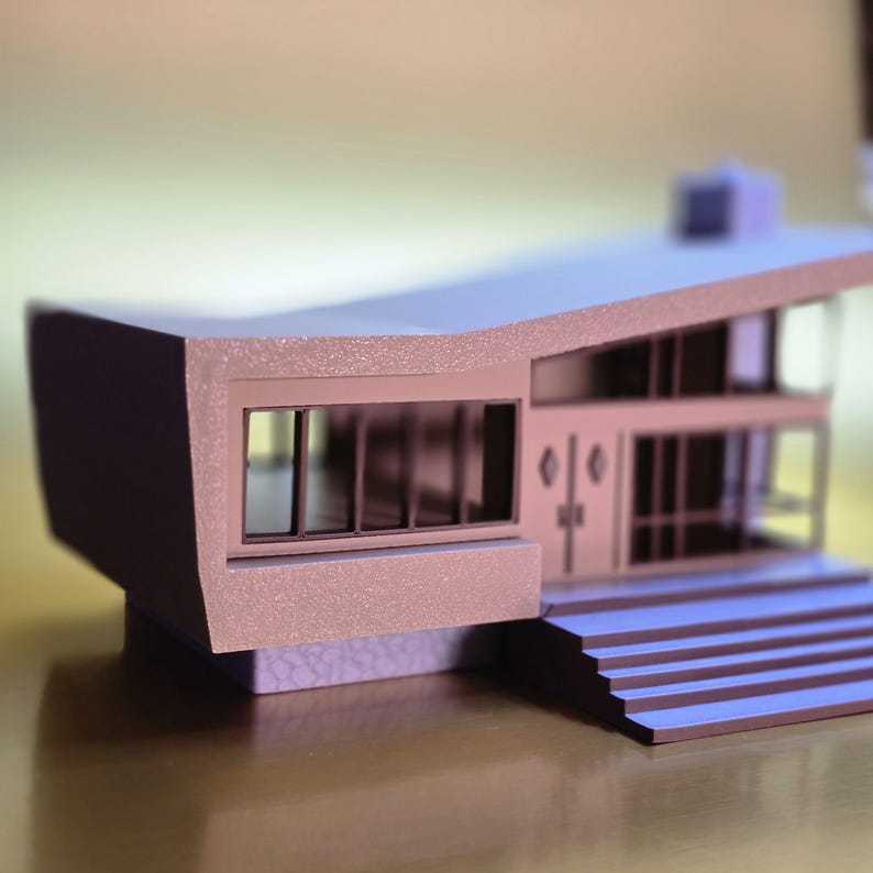 May include: A small, light brown model of a modern house with a flat roof and a set of stairs leading up to the front door. The house has large windows and a textured exterior.