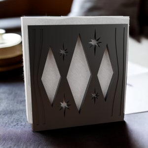 May include: A square napkin holder featuring a dark charcoal gray front panel with a laser-cut design. The pattern includes three vertical diamond shapes flanked by two curved lines, accented by four eight-pointed starburst motifs throughout.