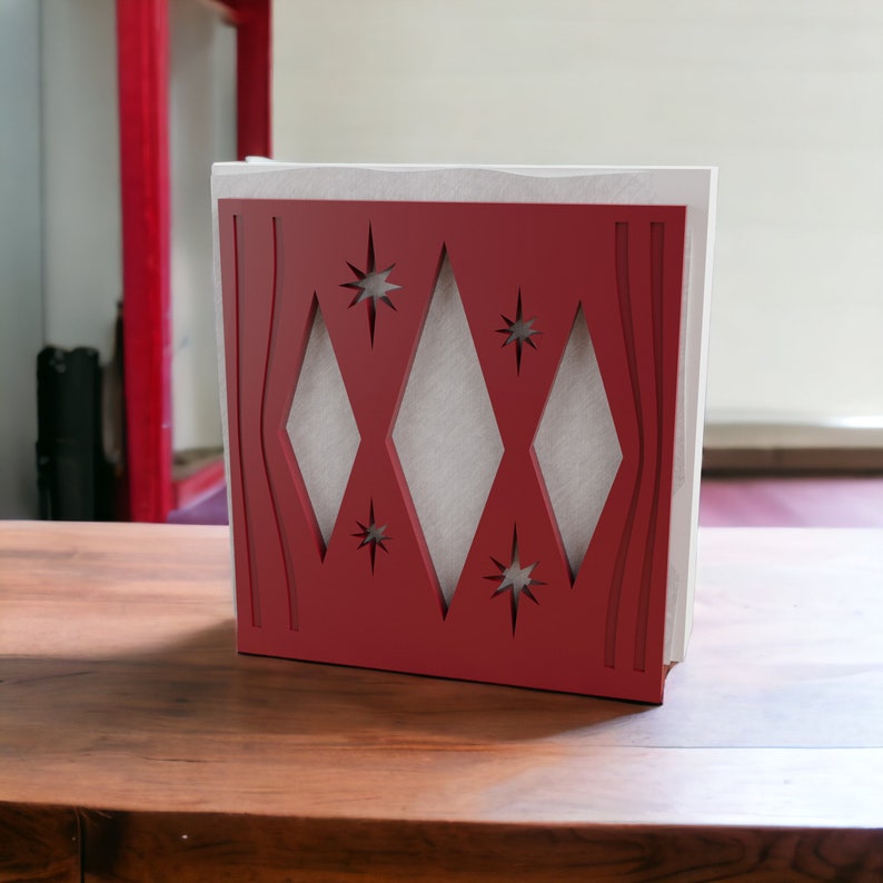 May include: A square napkin holder featuring a deep red front panel with cutouts in the shape of three large diamonds and four small starbursts, revealing a white interior structure. The design includes vertical curved lines on the sides.