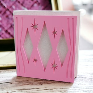 May include: A square pink napkin holder featuring a cutout design of three vertical diamonds, four-pointed starbursts, and curved vertical lines. The holder is constructed from a solid pink material with a white interior panel visible through the cutouts.