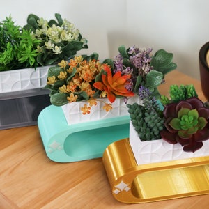 May include: Three different sized, oval-shaped planters with white geometric designs. The planters are made of a smooth, glossy material and are painted in gray, teal, and gold. Each planter holds a different type of plant, including a green leafy plant, a yellow and orange flowering plant, and a succulent.