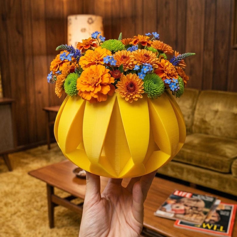 May include: A bright yellow, geometric vase filled with a vibrant arrangement of orange, green, and blue flowers. The vase has a unique, pleated design. The flowers are a mix of zinnias and other blooms, creating a colorful floral display.