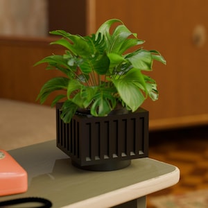May include: A green leafy plant in a black planter with vertical slats. The planter has a round base and sits on a wooden table.