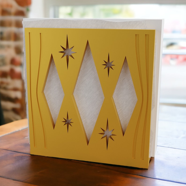 May include: A yellow napkin holder featuring a cut-out design of three vertical diamonds and four starburst shapes. The holder is constructed with a flat base and holds a stack of white paper napkins visible through the geometric cut-outs.