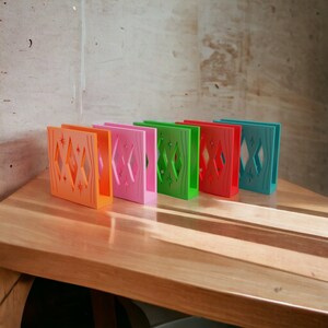 May include: Five rectangular napkin holders in orange, pink, green, red, and teal. Each holder features a cut-out design with diamond shapes and starburst accents on the side panels, standing upright in a row on a wooden surface.