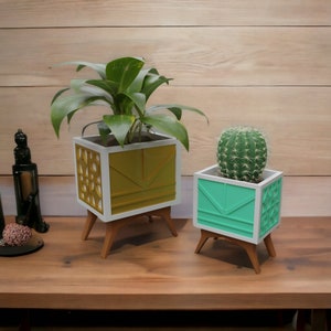 May include: Two geometric planter boxes with wooden legs. The larger planter is gold and white with a plant inside. The smaller planter is teal and white with a cactus inside.