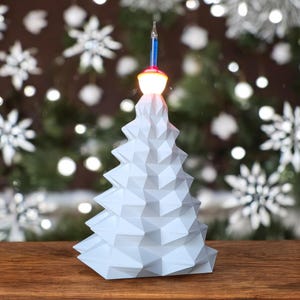 May include: A white, geometric, origami-style Christmas tree with a lit, clear, glass bulb on top. The tree is sitting on a wooden surface.