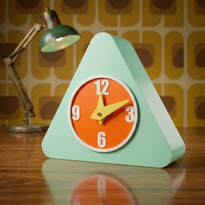 May include: A mint green triangular clock with an orange clock face. The clock face has white numbers and yellow hands. The clock is on a wooden surface. A green desk lamp is in the background.