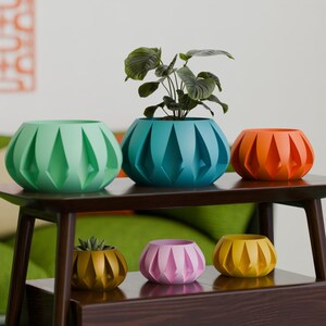 May include: A collection of colorful, geometric planters in shades of green, teal, orange, pink, and yellow. The planters are arranged on a dark wooden table, with a plant in the teal planter. The planters have a unique, faceted design.