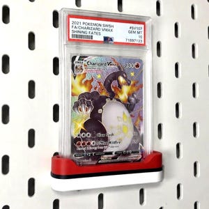 Könnte beinhalten: Eine PSA-bewertete 2021 Pokemon SWSH Charizard VMAX Shining Fates Sammelkarte in einer durchsichtigen Kunststoffhülle, präsentiert in einem rot-weißen Halter. Die Karte zeigt ein schwarzes Glurak mit Flammen und den Text 330 HP.