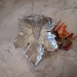 May include: A silver grape leaf dish with a cluster of grapes in the center. The dish is made of metal and has a detailed design.