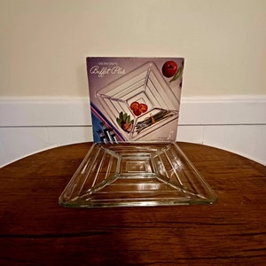 May include: A clear glass square serving dish with a raised, layered design. The dish is on a wooden table and the packaging is visible behind it. The packaging says "Colony Crafts Buffet Plus".