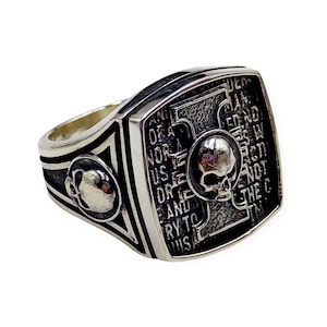 May include: A silver ring with a square face featuring a skull and crossbones design. The face of the ring is engraved with the text "ANTI-DEATH NOR A USELESS OR DRE AND TRY TO WIS", and the band of the ring is decorated with a skull design.