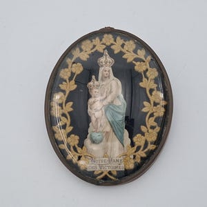 May include: An oval-shaped religious plaque featuring a central figure of a crowned woman holding a child, both wearing crowns. The figures are set against a black background, framed by a gold-colored floral wreath, with the text NOTRE DAME DES VICTOIRES.