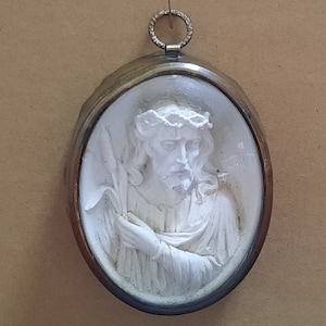 Puede incluir: Un camafeo ovalado de cerámica blanca con Jesús Cristo con una corona de espinas. El camafeo está enmarcado en un marco de metal plateado y cuelga de una cadena plateada.