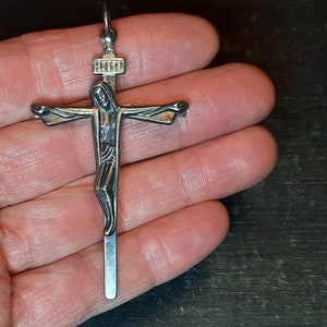 Może przedstawiać: Srebrny wisiorek z krzyżem z detaliczną figurą Jezusa Chrystusa. Wisiorek ma prosty wzór krzyża z małym napisem u góry.