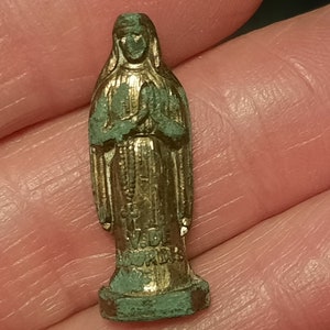 以下が含まれることがあります： 聖母マリアの小さな金色の金属製の置物。置物は、祈りを捧げるために手を合わせ、立っています。置物は緑色の緑青がかかっています。