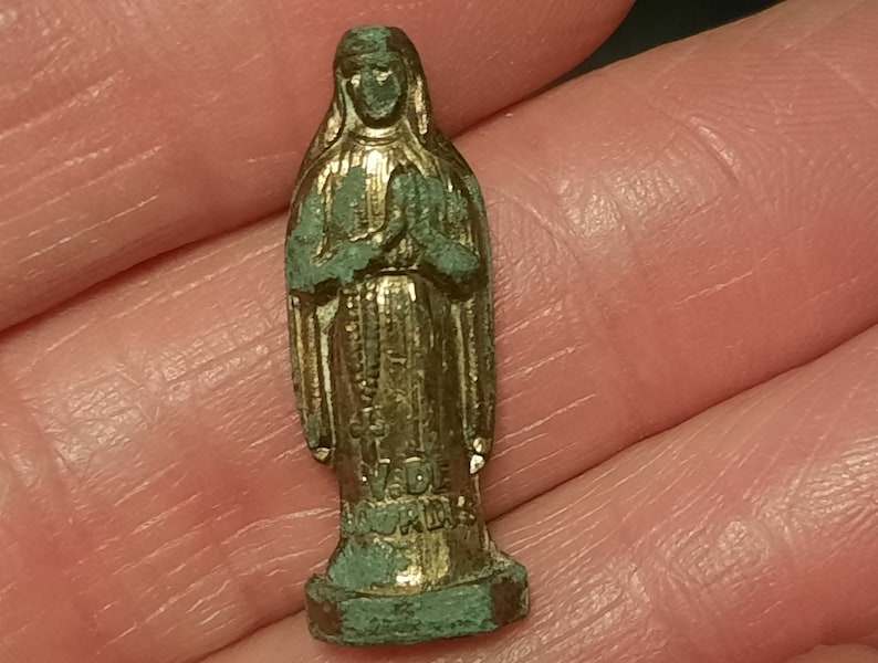 以下が含まれることがあります： 聖母マリアの小さな金色の金属製の置物。置物は、祈りを捧げるために手を合わせ、立っています。置物は緑色の緑青がかかっています。
