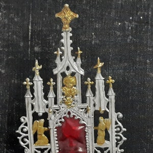 May include: A silver and gold metal religious ornament with a red glass center piece. The ornament is shaped like a church with a pointed roof and multiple spires.