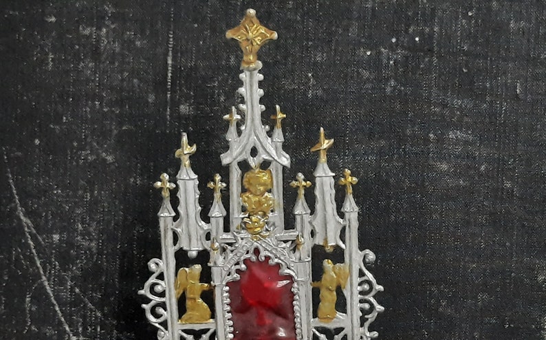 May include: A silver and gold metal religious ornament with a red glass center piece. The ornament is shaped like a church with a pointed roof and multiple spires.