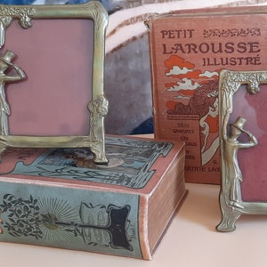 May include: Two antique brass picture frames with ornate designs and a figure of a person in a top hat on each side. The frames are sitting on top of a book titled "De Meuwe" with a green and blue cover.