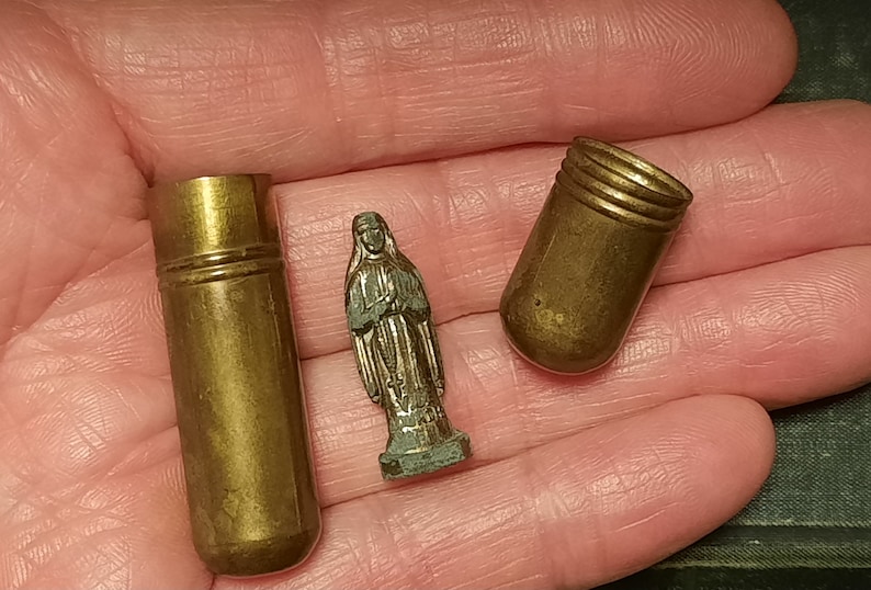 以下が含まれることがあります： 金色の小さな金属製の容器が3つあります。1つの容器は開いていて、他の2つは閉じています。開いている容器は円筒形で、丸みを帯びた上部があります。閉まっている容器は円筒形で、丸みを帯びた上部と底部があります。中央には、銀色の小さな金属製の女性の像があります。
