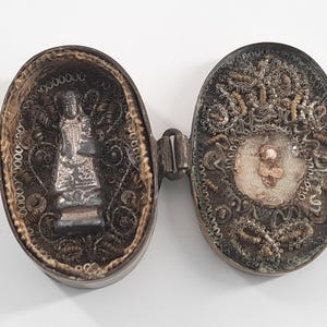 May include: An open, antique oval reliquary with a hinged design. One side features a silver statue, the other a detailed embroidered image. The reliquary is framed with intricate metalwork and decorative trim.