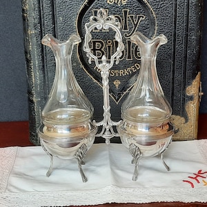 Puede incluir: Un juego de dos jarras de plata con botellas de vidrio transparente y un asa decorativa. El juego de jarras está sobre un paño de encaje blanco con la palabra "JHS" bordada en rojo.