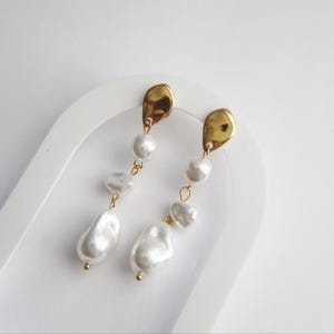 May include: A pair of elegant dangle earrings featuring irregular white pearls suspended from gold-colored, teardrop-shaped metal accents. The earrings have a delicate, feminine design, perfect for adding a touch of sophistication to any outfit.