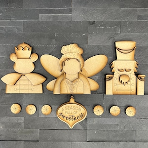 May include: Wooden cutout ornaments featuring a nutcracker, a fairy, and a prince. The ornaments are unfinished and ready to be painted. The ornaments are surrounded by small wooden candy cane cutouts. The ornament in the center has the text "A season full of sweetness".