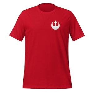 Puede incluir: Una camiseta roja de manga corta con un símbolo blanco de la Alianza Rebelde en el pecho izquierdo.