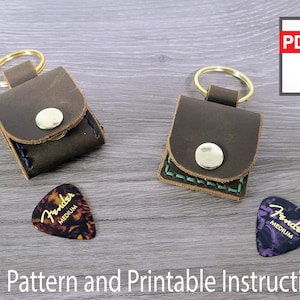May include: Two brown leather guitar pick holders with a snap closure and a key ring. The holders are on a wooden surface with two guitar picks, one brown and one purple.