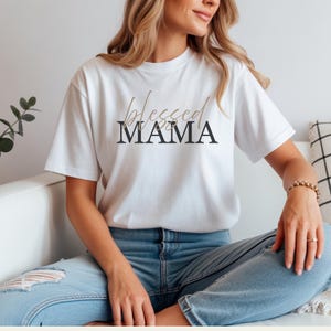 May include: A white short-sleeved t-shirt featuring the text "blessed" in a light brown cursive script above the word "MAMA" in a bold, black, uppercase serif font. The shirt is paired with light blue denim jeans that have a distressed knee detail.