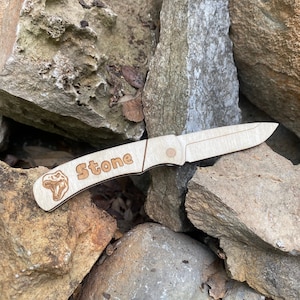 May include: A wooden pocket knife with a dinosaur design on the handle. The knife is engraved with the name "Stone".