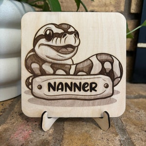 May include: A square wooden plaque with a cartoon snake design. The snake is brown and white, with the name "NANNER" engraved on a banner. The plaque is displayed on a wooden stand.