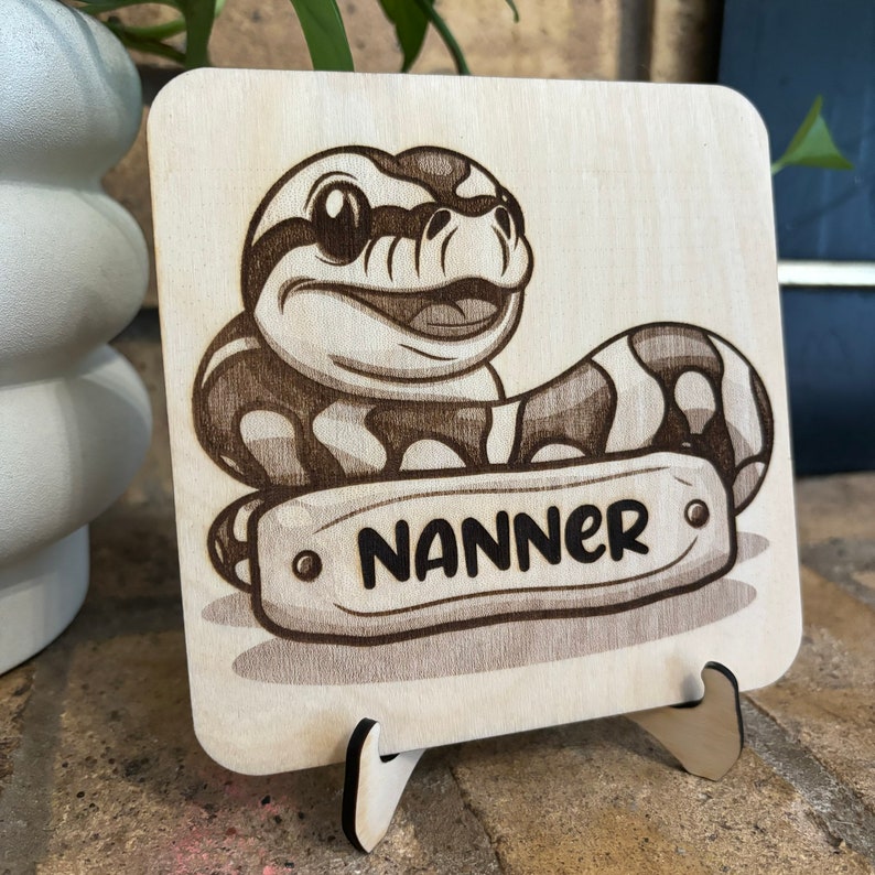 May include: Wooden square plaque featuring a cartoon snake with the name "NANNER" on a banner. The snake is brown and white, with a smiling face. The plaque is displayed on a wooden stand.