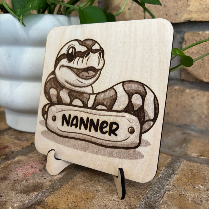 May include: A wooden square with a cartoon snake illustration. The snake is smiling and has a patterned body. The name "NANNER" is on a banner. The wooden piece is displayed on a small wooden stand.