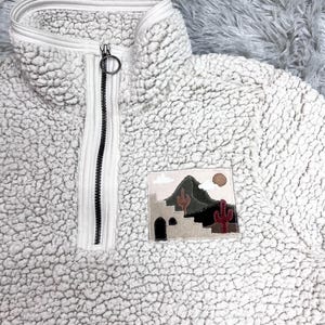 May include: A white sherpa fleece pullover with a half-zip closure and a colorful embroidered patch depicting a desert landscape with a mountain, cacti, and a building.