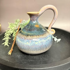 May include: A small, handcrafted ceramic pitcher with a curved handle. The pitcher features a blend of blue, green, and brown glaze, creating a unique, marbled effect. The handle and rim have a lighter, tan color. A decorative piece for the home.