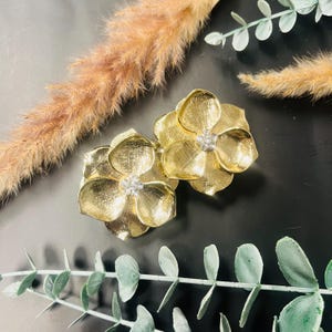 May include: A pair of gold-coloured flower-shaped earrings with a textured finish. Each earring has a small, clear crystal in the centre. The earrings are set against a dark background with decorative foliage.