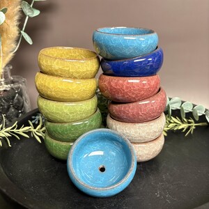 May include: A stack of small, colorful ceramic bowls in shades of yellow, green, blue, pink, and beige. Each bowl has a cracked glaze finish. The bowls are stacked on a black tray, with a single bowl in the foreground.